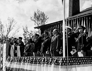 Мужчины в костюмах и военной форме на помосте, украшенном флагами США, отдают честь